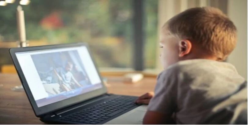 A small boy is watching a video on his laptop in a blurry background.